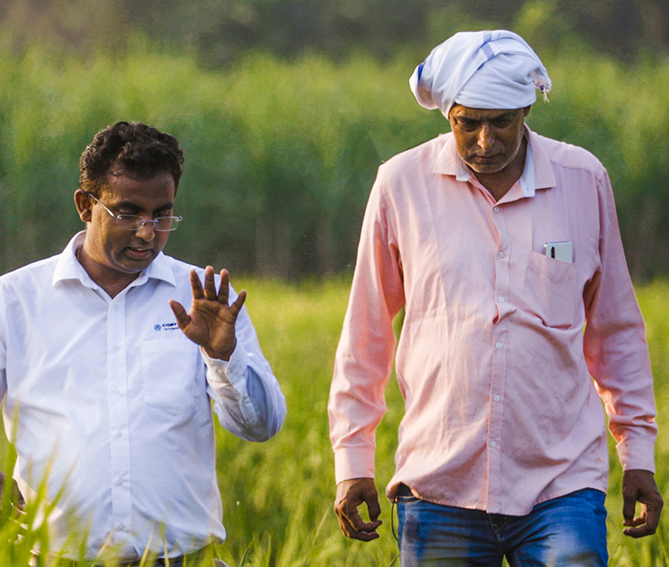 Two men walking through a green agricultural field, having a discussion; one is wearing a white shirt and glasses, the other in a pink shirt with a head wrap.