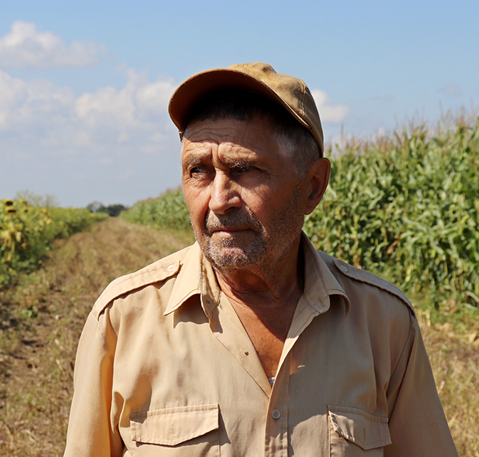 Elderly male farmer wearing a beige shirt and cap standing in a field with tall crops under a clear sky.