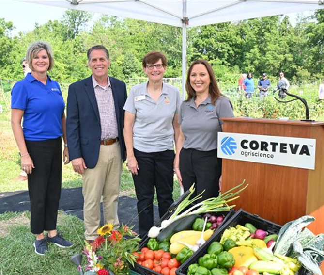 Four people standing under a white tent next to a podium with a Corteva Agriscience sign, with a display of fresh vegetables in front of them at an outdoor event.