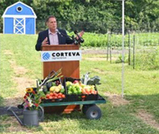 Man speaking at a podium with a Corteva Agriscience sign, outdoors near a field, with fresh vegetables and flowers displayed on a cart in front of him and a blue barn in the background.