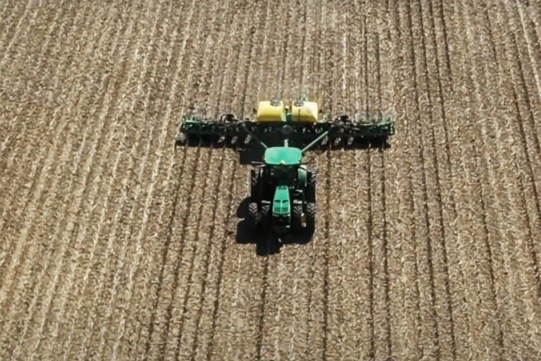 Aerial view of a green tractor plowing a harvested field with farming equipment attached.