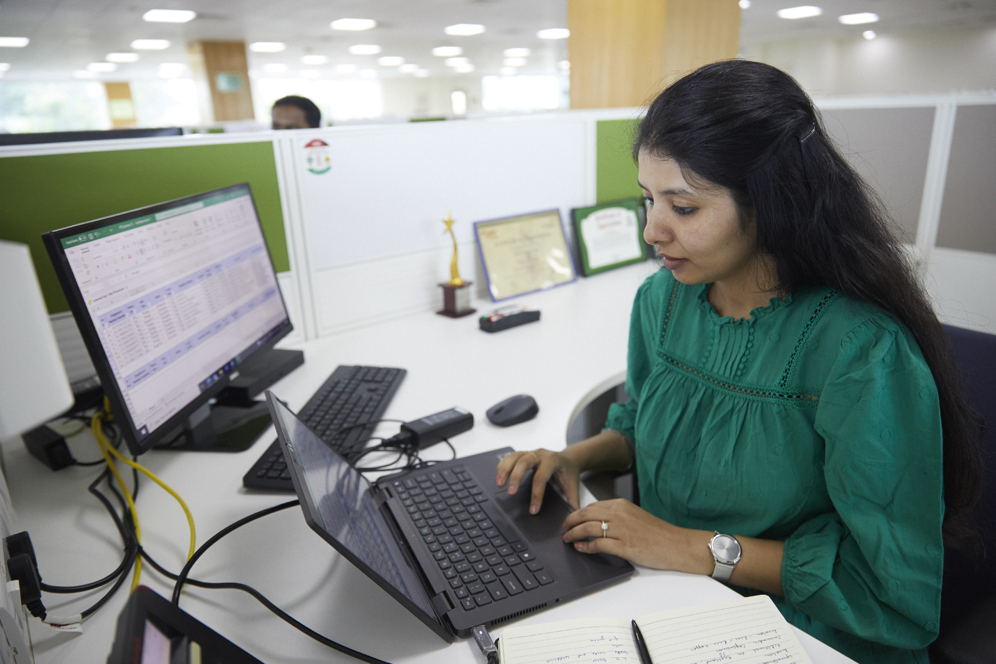 Woman working at her desk in an office, using a laptop and external monitor displaying spreadsheets, with certificates and a trophy in the background.