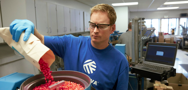 Laboratory technician wearing gloves and safety glasses pouring red coated seeds from a paper bag into a testing machine in a lab setting.