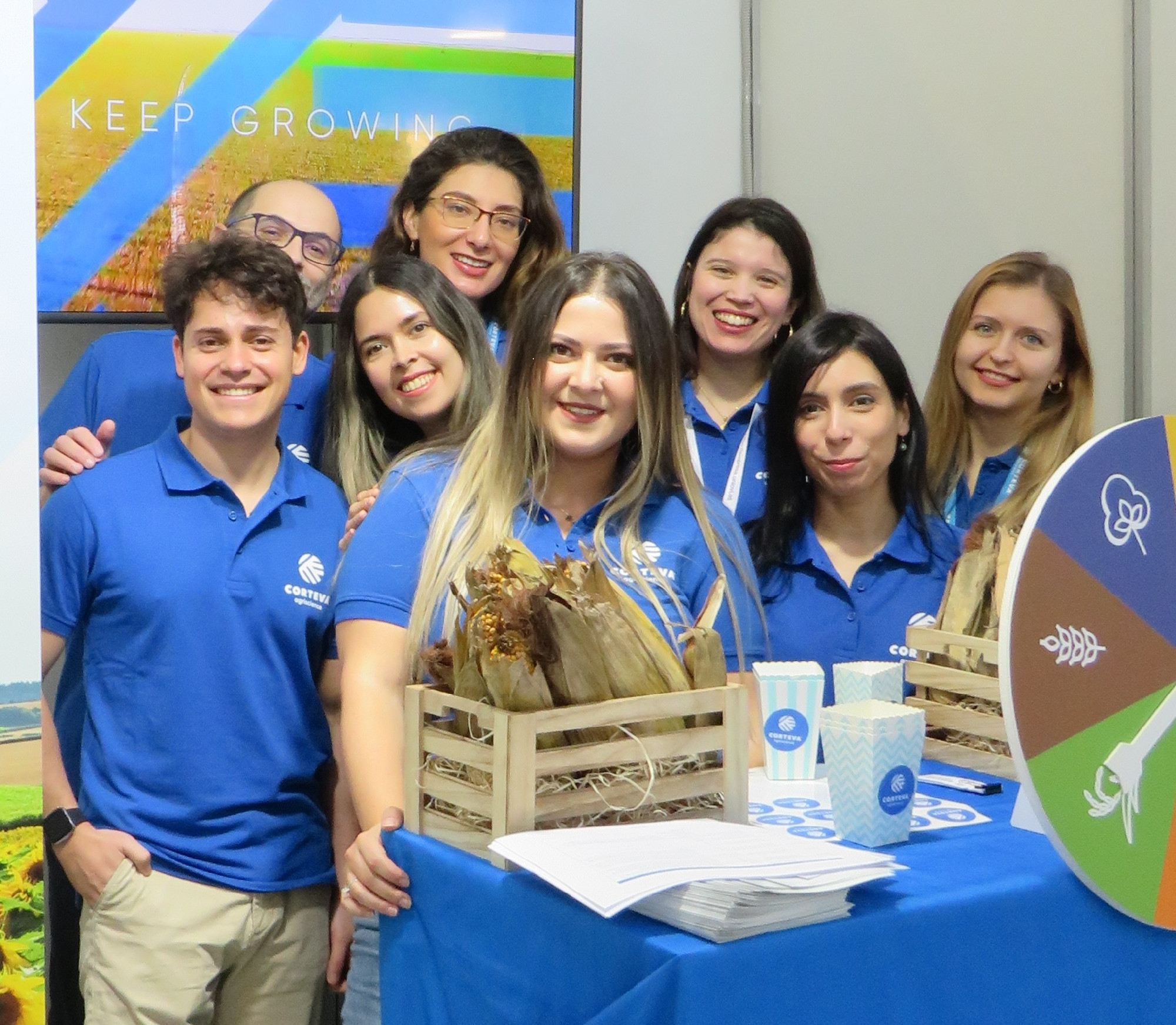 Group of smiling team members wearing blue Corteva shirts standing with a display of informational materials and display crates of dried corn.